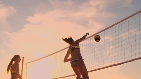 SLOW-MOTION-LOW-ANGLE-CLOSE-UP-LENS-FLARE:-Young-female-volleyball-players-pass-and-spike-the-ball-over-the-net-on-a-sunny-summer-evening.-Fit-Caucasian-girls-playing-beach-volleyball-at-sunset.